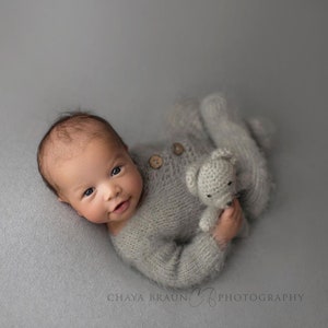 Könnte beinhalten: Ein Baby trägt einen grauen Strick-Strampler mit einem gehäkelten Bären. Das Baby liegt auf einem grauen Hintergrund und schaut in die Kamera.