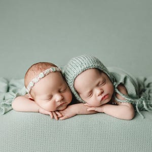 Op de afbeelding: Twee pasgeboren baby's slapen op een lichtgroen, getextureerd deken. Eén baby draagt ​​een lichtgroene gebreide muts en de andere baby draagt ​​een wit hoofdband met kleine bloemen.