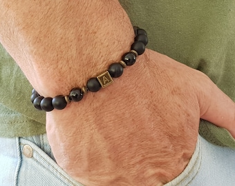 May include: Black beaded bracelet with a gold-colored square charm featuring the letter "A". The bracelet is made of matte black beads and is accented with gold-colored spacers. A stylish accessory.
