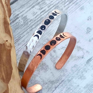 May include: Two metal cuff bracelets with a moon phase design. One bracelet is silver with a hammered texture, and the other is copper. The black moon phase graphics are evenly spaced along the bracelets. The bracelets are displayed on a light wood surface.