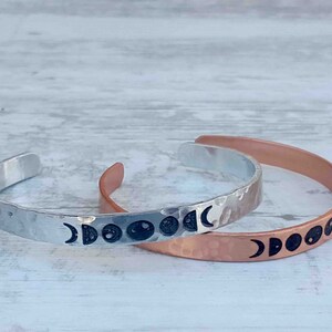 May include: Two metal cuff bracelets, one silver and one copper, each featuring a hammered texture. The silver bracelet displays the phases of the moon in black, while the copper bracelet also showcases the moon phases.
