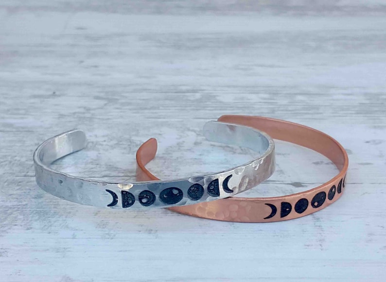 May include: Two metal cuff bracelets, one silver and one copper, each featuring a hammered texture. The silver bracelet displays the phases of the moon in black, while the copper bracelet also showcases the moon phases.
