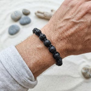 May include: A black lava stone bead bracelet featuring a series of porous, dark volcanic rock spheres strung together with a durable black cord. The bracelet is designed with a knotted pattern that secures each individual bead in a fixed position.