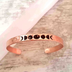 May include: A copper-colored cuff bracelet with a hammered texture. The bracelet features a series of black moon phase illustrations across the top. The open-ended design allows for easy wear. The bracelet is displayed on a light-colored surface.