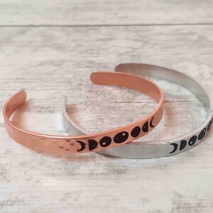 May include: Two open cuff bracelets. One is copper-colored with a hammered texture and black moon phase design. The other is silver-colored with a smooth finish and a black moon phase design. Both are on a light wood-grain surface.