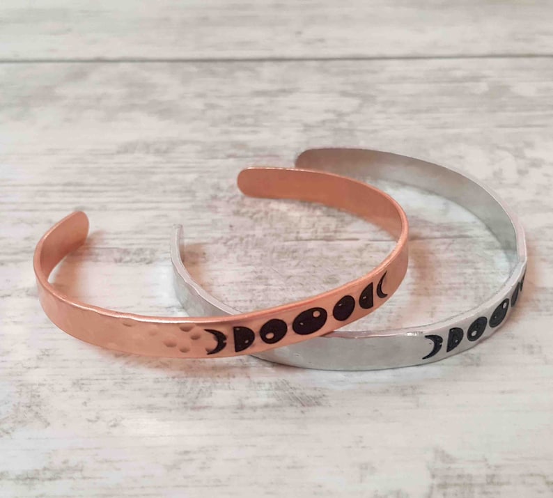 May include: Two open cuff bracelets. One is copper-colored with a hammered texture and black moon phase design. The other is silver-colored with a smooth finish and a black moon phase design. Both are on a light wood-grain surface.