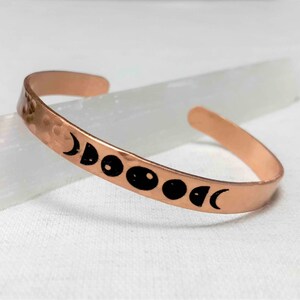 May include: A copper cuff bracelet with a hammered texture. The bracelet features a series of black moon phase designs. The open-ended bracelet is displayed on a white surface.