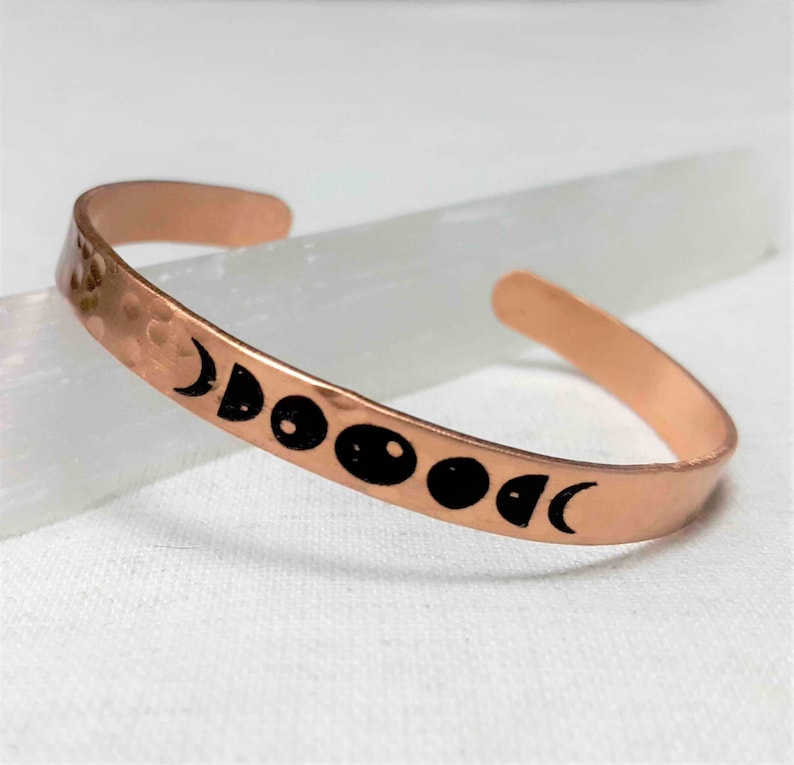 May include: A copper cuff bracelet with a hammered texture. The bracelet features a series of black moon phase designs. The open-ended bracelet is displayed on a white surface.