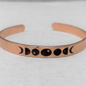 May include: A copper-colored cuff bracelet with a series of black moon phase symbols. The open-ended bracelet is made of a thin, flat metal band. The moon phase symbols are evenly spaced across the front of the bracelet.