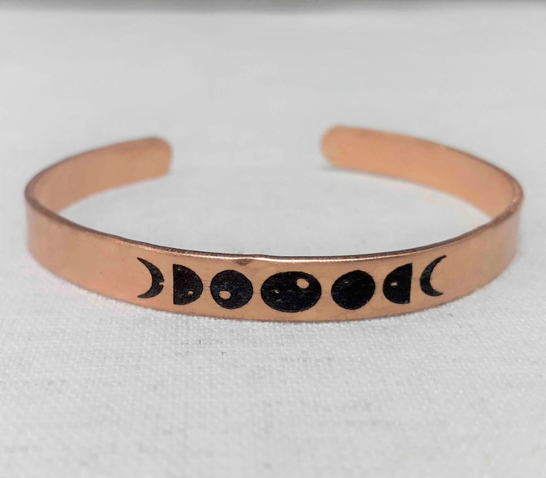 May include: A copper-colored cuff bracelet with a series of black moon phase symbols. The open-ended bracelet is made of a thin, flat metal band. The moon phase symbols are evenly spaced across the front of the bracelet.