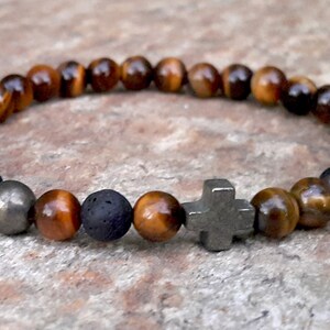 May include: A beaded bracelet featuring tiger's eye, lava stone, and pyrite beads. The bracelet includes a silver-colored cross charm. The beads are brown and gold, and the bracelet is displayed on a stone surface.