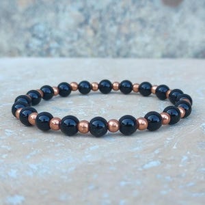 Puede incluir: Un brazalete de cuentas negras y cobre. El brazalete está hecho con cuentas negras y cobre alternadas.