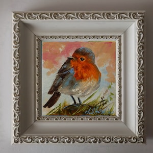 May include: An oil painting of a robin with a bright orange breast and grey-brown feathers, perched on green foliage against a soft pink and white sky. The artwork is encased in a square, off-white frame with an ornate, swirling relief pattern.