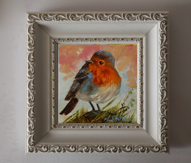 May include: An oil painting of a robin with a bright orange breast and grey-brown feathers, perched on green foliage against a soft pink and white sky. The artwork is encased in a square, off-white frame with an ornate, swirling relief pattern.