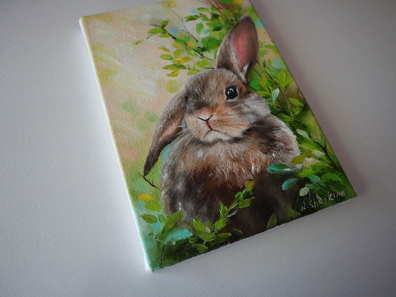 May include: A rectangular canvas painting featuring a brown rabbit with one upright ear and one floppy ear, nestled among vibrant green foliage. The background displays soft yellow and light green tones, with the artist's signature visible in the corner.