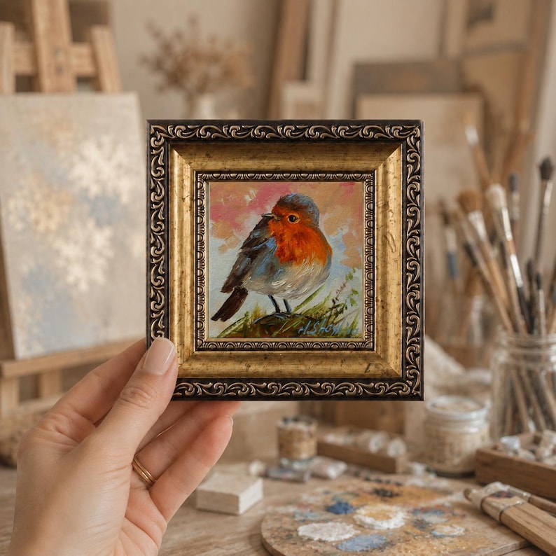 May include: A small square oil painting of a robin with a bright orange breast, perched on a branch against a soft pink and blue background. The artwork is encased in an ornate, dark gold-toned frame with a detailed, embossed scrollwork pattern.