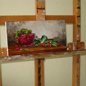 May include: An oil painting on canvas depicting a glass bowl filled with bright red raspberries and green leaves resting on a dark wooden surface. The composition features scattered berries and foliage in the foreground against a neutral gray background.