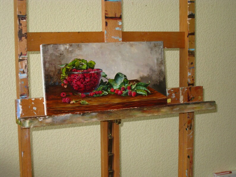 May include: An oil painting on canvas depicting a glass bowl filled with bright red raspberries and green leaves resting on a dark wooden surface. The composition features scattered berries and foliage in the foreground against a neutral gray background.