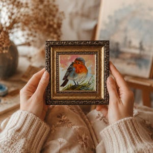 May include: A small square painting of a robin with a bright orange breast and grey wings, set against a soft pink and white background. The artwork is encased in an ornate, antique-style gold frame with a textured, dark metallic finish.