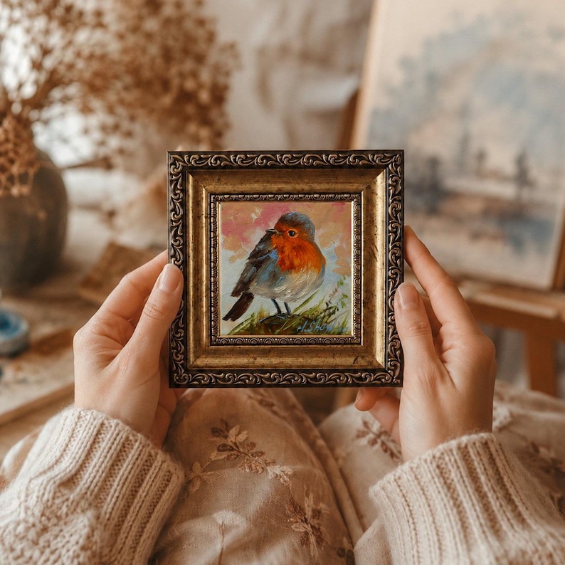 May include: A small square painting of a robin with a bright orange breast and grey wings, set against a soft pink and white background. The artwork is encased in an ornate, antique-style gold frame with a textured, dark metallic finish.