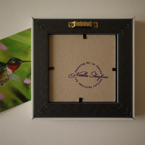 May include: The back of a square picture frame with a dark brown finish, featuring a gold-toned metal sawtooth hanger. A circular purple stamp on the cardboard backing reads Painting Oil on Canvas Natalia Shaykina Gallery Paintings Art.