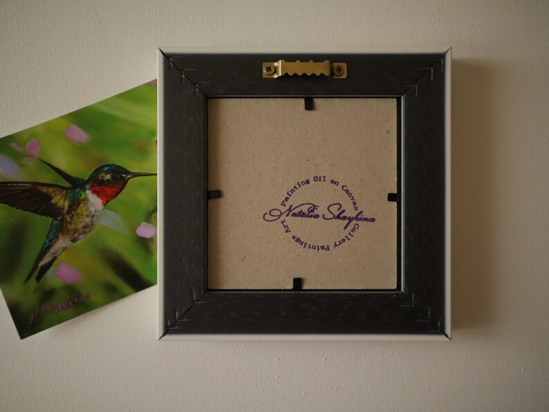 May include: The back of a square picture frame with a dark brown finish, featuring a gold-toned metal sawtooth hanger. A circular purple stamp on the cardboard backing reads Painting Oil on Canvas Natalia Shaykina Gallery Paintings Art.