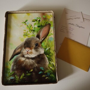 May include: A rectangular painting of a brown rabbit nestled in green foliage, presented in a cardboard box. Beside the box lies a certificate of authenticity signed by Natalia Shaykina and a small, plain, rectangular gold-colored envelope.