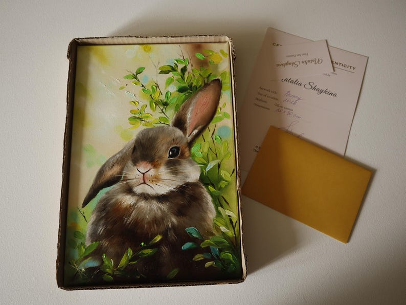 May include: A rectangular painting of a brown rabbit nestled in green foliage, presented in a cardboard box. Beside the box lies a certificate of authenticity signed by Natalia Shaykina and a small, plain, rectangular gold-colored envelope.