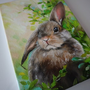 May include: An oil painting on canvas depicting a brown rabbit with one upright ear and one folded ear, positioned among bright green foliage. The background features soft yellow and light green tones, with the artist's signature visible in the corner.