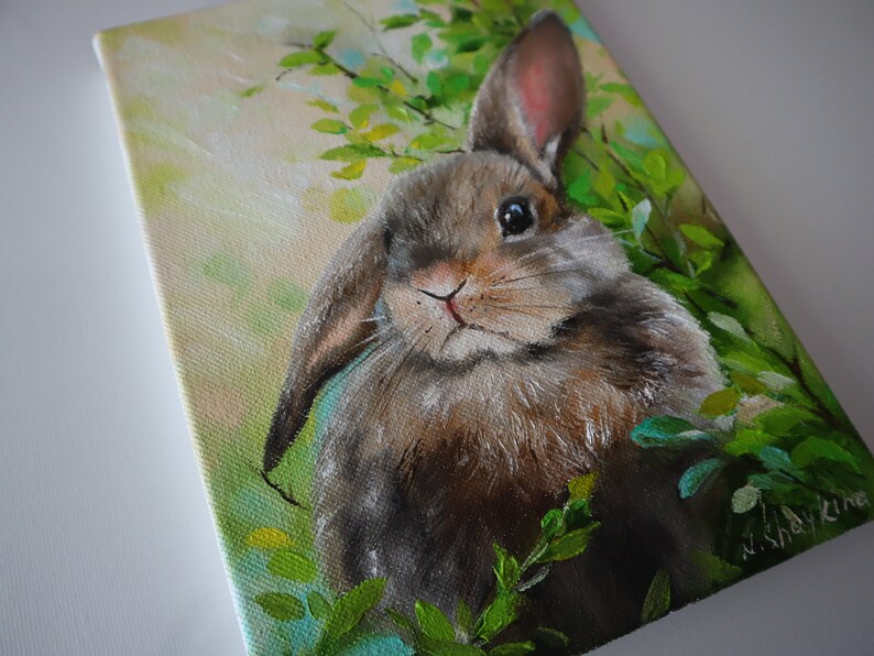 May include: An oil painting on canvas depicting a brown rabbit with one upright ear and one folded ear, positioned among bright green foliage. The background features soft yellow and light green tones, with the artist's signature visible in the corner.