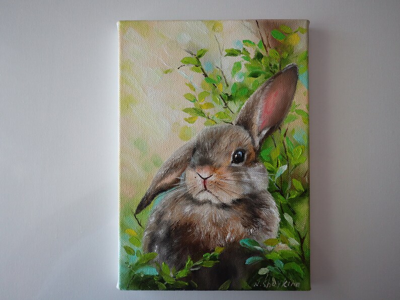 May include: An oil painting on canvas depicting a brown rabbit with one upright ear and one floppy ear, nestled among vibrant green foliage. The background features soft, light-filled brushstrokes in shades of yellow and cream, with the artist's signature N. Shaykina in the bottom right corner.