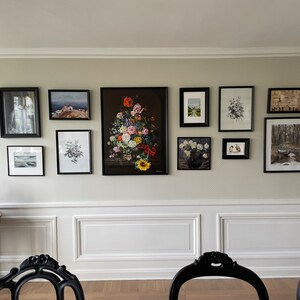 May include: A gallery wall featuring framed art prints of various sizes and styles. The collection includes landscape paintings, floral arrangements, and abstract images, all in black frames. The arrangement is displayed on a neutral-colored wall.