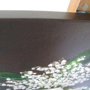 May include: A painting of a bouquet of white lily of the valley flowers with green leaves against a dark brown background. The painting is on a wooden frame with a dark brown border.