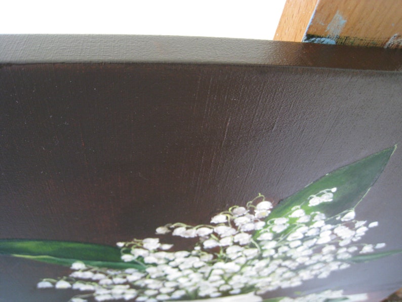 May include: A painting of a bouquet of white lily of the valley flowers with green leaves against a dark brown background. The painting is on a wooden frame with a dark brown border.