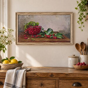 May include: A rectangular framed oil painting depicting a glass bowl filled with bright red raspberries and green leaves resting on a dark wooden surface. The artwork features a neutral gray background and is displayed in a light-colored wooden frame.