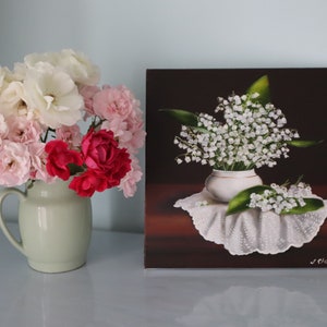 May include: A still life painting and a vase of flowers. The painting depicts white lily of the valley flowers in a white vase on a white lace cloth. A vase of pink, white, and red roses sits to the left.