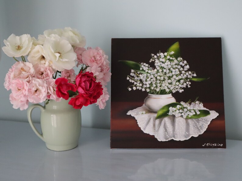 May include: A still life painting and a vase of flowers. The painting depicts white lily of the valley flowers in a white vase on a white lace cloth. A vase of pink, white, and red roses sits to the left.