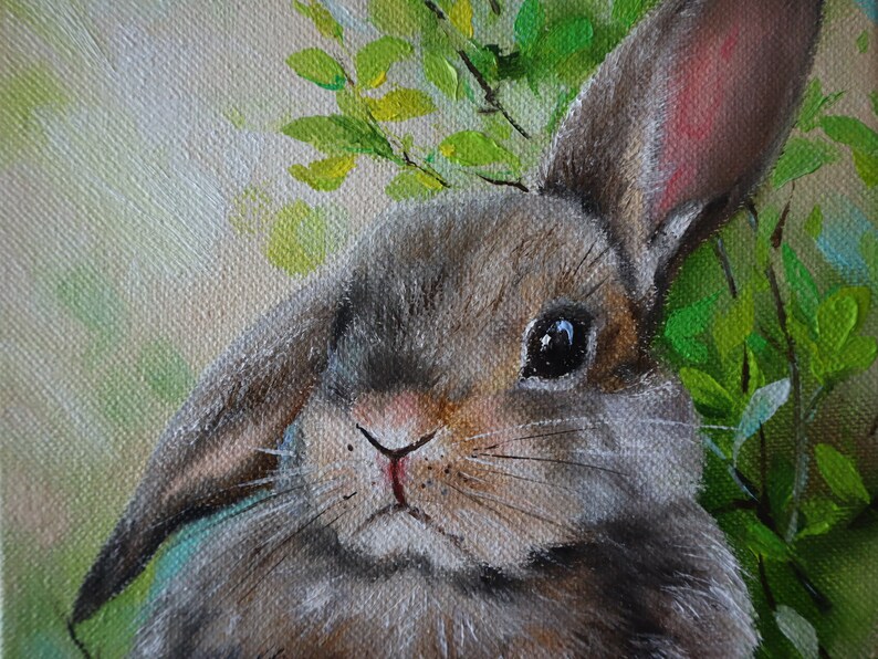 May include: An oil painting on canvas depicting a brown rabbit with one upright ear and one folded ear. The rabbit is positioned against a backdrop of vibrant green leaves and soft, light-colored foliage, rendered with visible brushstrokes.