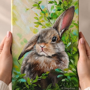 May include: An oil painting on canvas depicting a brown rabbit with one upright ear and one folded ear, nestled among vibrant green foliage. The background features soft yellow and light green brushstrokes, with the artist signature N.Shaykina.