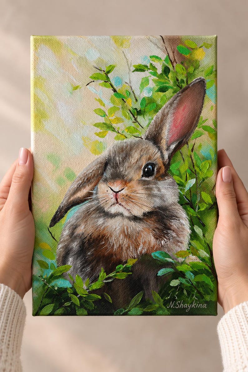May include: An oil painting on canvas depicting a brown rabbit with one upright ear and one folded ear, nestled among vibrant green foliage. The background features soft yellow and light green brushstrokes, with the artist signature N.Shaykina.