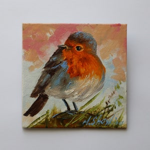 May include: An oil painting on a square canvas depicting a European robin with a bright orange breast, grey-brown wings, and a dark eye. The bird is perched on green grass against a soft, textured background of muted pink and light blue brushstrokes.