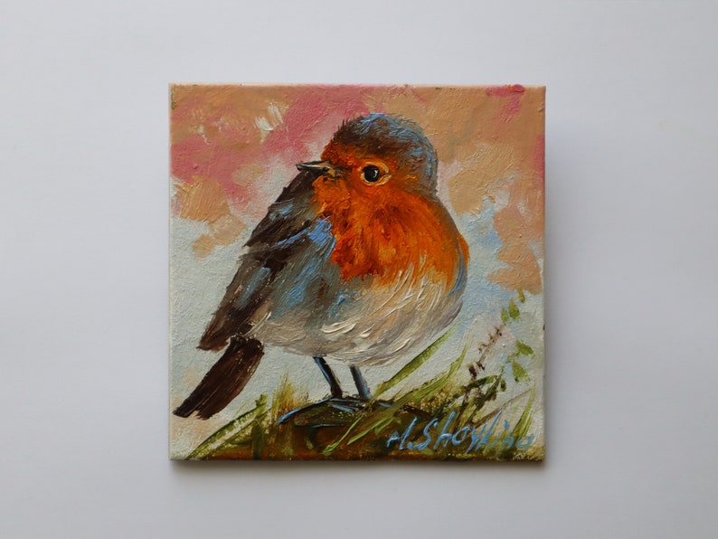 May include: An oil painting on a square canvas depicting a European robin with a bright orange breast, grey-brown wings, and a dark eye. The bird is perched on green grass against a soft, textured background of muted pink and light blue brushstrokes.