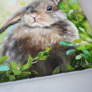 May include: An oil painting on canvas depicting a brown lop-eared rabbit nestled among vibrant green foliage. The rabbit has soft fur, dark eyes, and long ears. The artist's signature, N. Shaykina, is visible in the bottom right corner of the canvas.