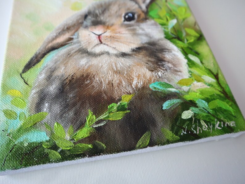 May include: An oil painting on canvas depicting a brown lop-eared rabbit nestled among vibrant green foliage. The rabbit has soft fur, dark eyes, and long ears. The artist's signature, N. Shaykina, is visible in the bottom right corner of the canvas.