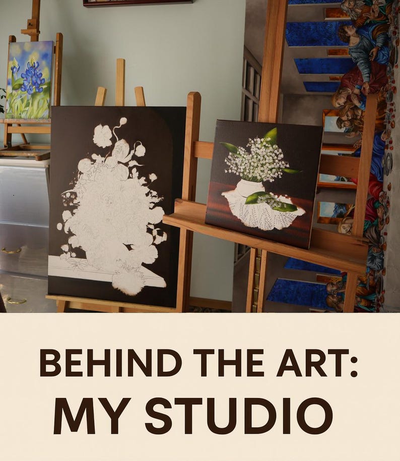 May include: Three wooden easels holding canvases in a studio. One canvas shows a blue iris painting, another features a black and white floral outline, and the third displays a realistic painting of white lily of the valley flowers in a vase.