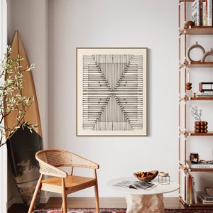 May include: A framed abstract art print with a geometric design in black lines on a cream background hangs on a white wall. A wooden chair with a woven back and a round coffee table with a stone base are in front of a patterned rug.