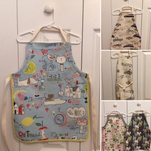 May include: Four children's aprons with different colorful patterns. The aprons are hanging on white hangers. The patterns include a blue background with a dog, cat, bird, and house, a beige background with cars, a beige background with airplanes, and a beige background with owls and horses.