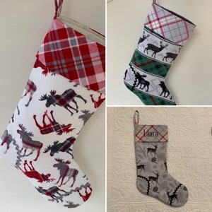 May include: Three Christmas stockings with a woodland theme. The first stocking is red and white plaid with a white background and moose silhouettes. The second stocking is green, white, and gray with a white background and moose and reindeer silhouettes. The third stocking is gray with a white background and a moose silhouette. The third stocking has the name "Laura" embroidered on the cuff.