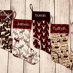 May include: Four personalized Christmas stockings with a woodland theme. The stockings are made of red and white plaid fabric with a cream-colored fabric featuring a pattern of moose, deer, and trees. The stockings are personalized with the names "Rachel", "James", "Susan", and "Nathan".