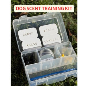 May include: A clear plastic dog scent training kit with the text "DOG SCENT TRAINING KIT". The kit contains small white labeled containers with the words "CLOVE", "BIRCH", "ANISE", and "CYPRESS". Also included are small bottles, a metal tin, and a blue pen.